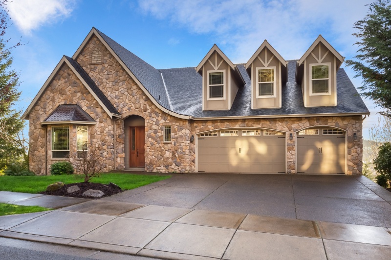 A stone-faced house with three dormer windows, a wooden front door, and a two-car garage. The driveway is wet, and the sky is partly cloudy with some blue visible. Green grass and trees surround the home.