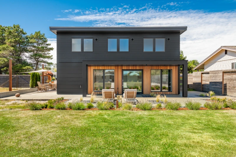 Modern two-story house with dark siding and large windows, wooden accents around the doors, a patio with two lounge chairs, and a landscaped yard with green grass and garden beds under a blue sky.